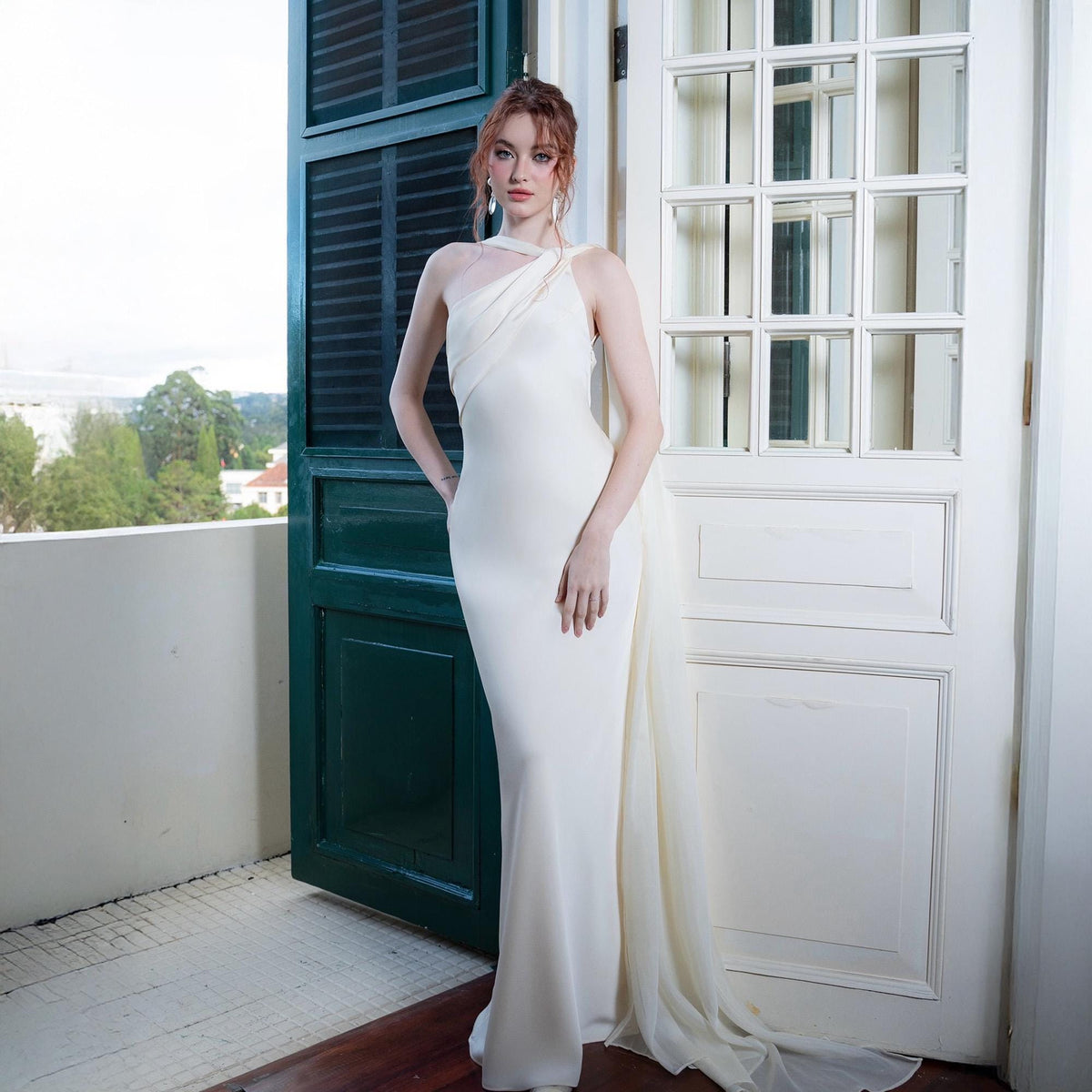 May include: A floor-length, ivory-colored gown with a draped, one-shoulder neckline and a flowing train. The dress has a fitted silhouette that flares slightly at the hem. The model is standing on a balcony with a green door and white door in the background.