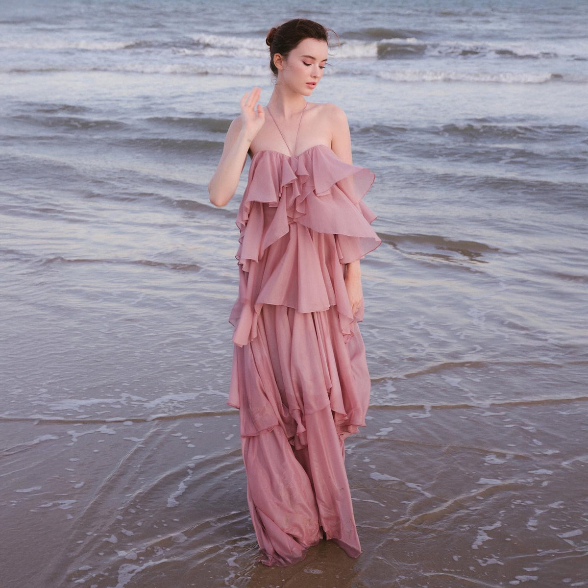 May include: A strapless, tiered, dusty rose maxi dress. The dress features multiple layers of ruffles, creating a flowing silhouette. The model is standing in the ocean, with the waves gently lapping at the hem of the dress. The dress is made of a soft, lightweight fabric.