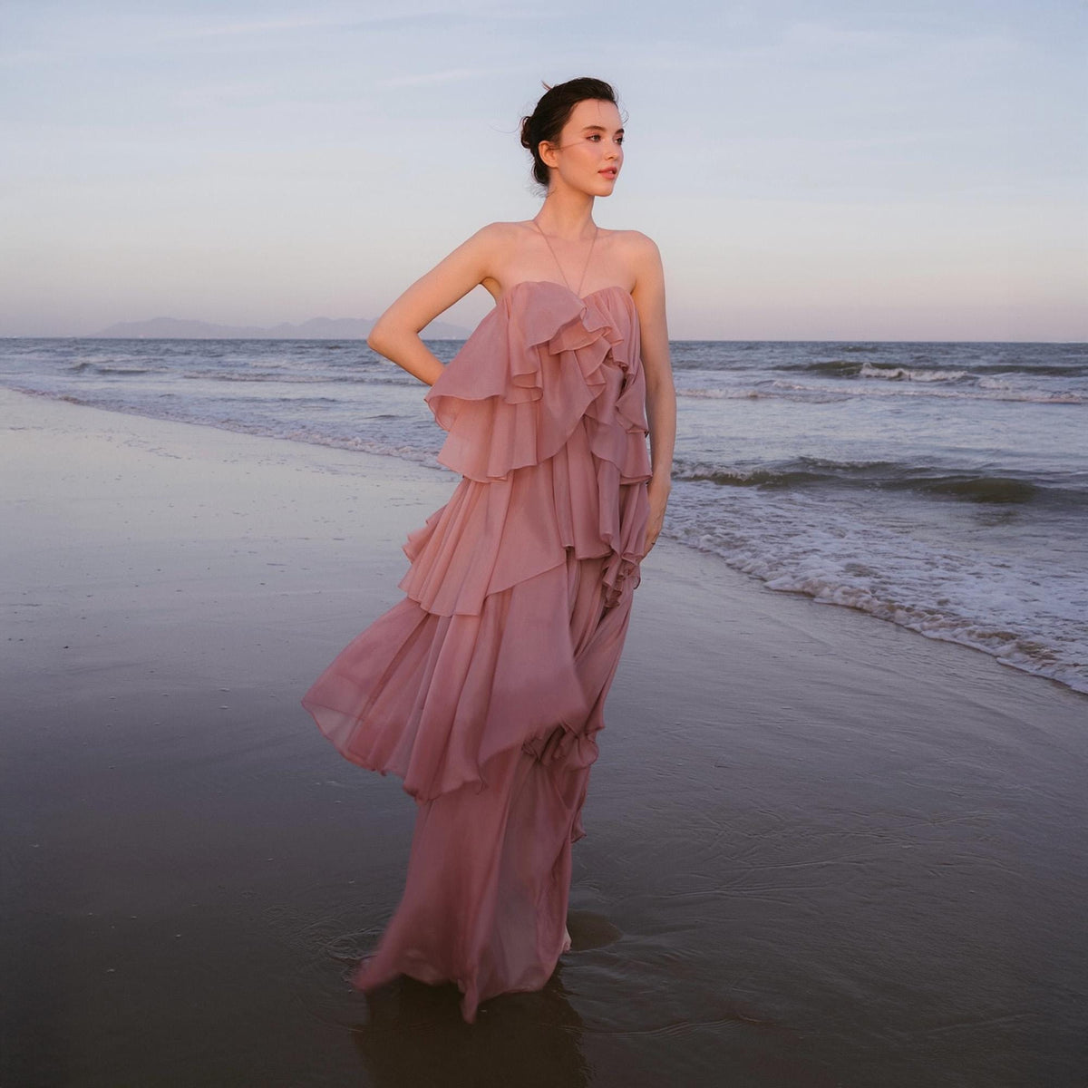 May include: A strapless, dusty rose-colored gown with layered ruffles. The dress is floor-length and flowing, with a soft, romantic aesthetic. The image is taken on a beach, with the ocean and sky in the background.