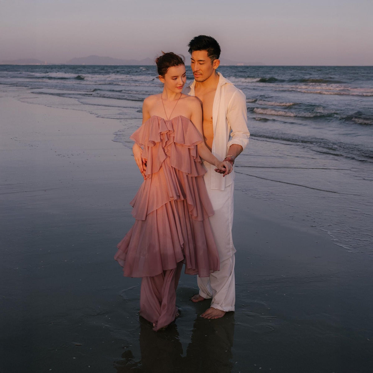 May include: A couple stands on a beach at sunset. The woman wears a layered, strapless, dusty rose gown. The man is in white pants and a white open shirt. The ocean and sky are in the background.
