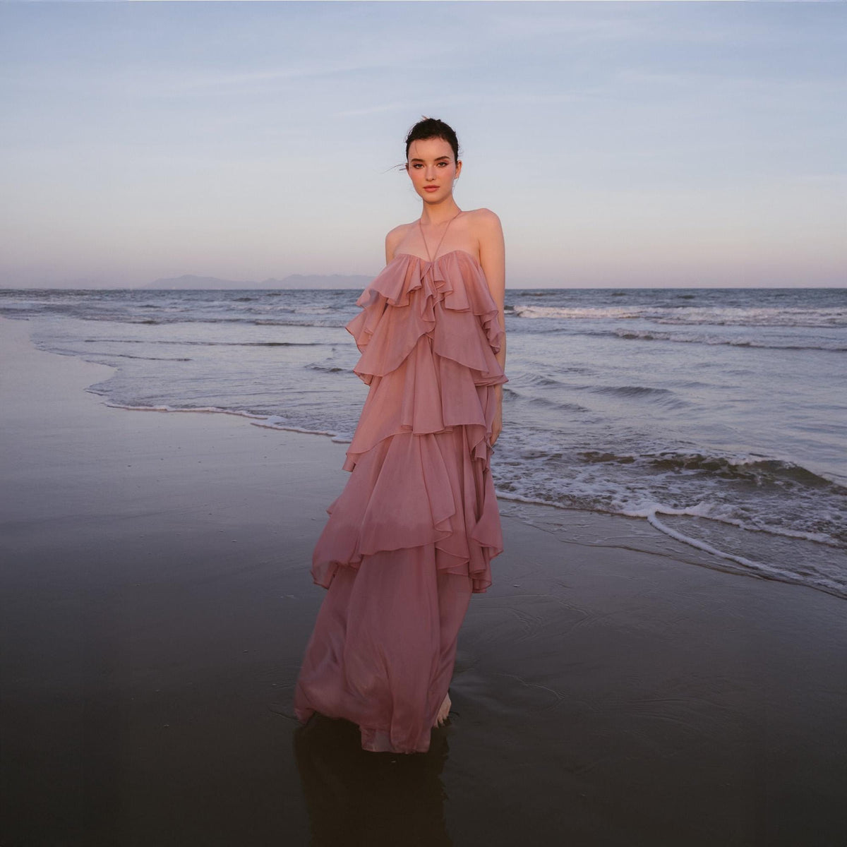 May include: A strapless, tiered, dusty rose-colored gown. The dress features multiple layers of flowing fabric, creating a ruffled effect. The model is standing on a beach with the ocean and sky in the background.