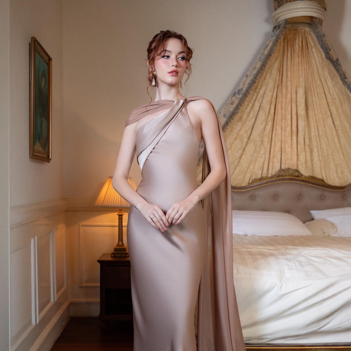 May include: A floor-length, champagne-colored gown with a draped shoulder detail and a high slit. The dress is made of a silky material and has a fitted silhouette. The model is standing in a bedroom setting.