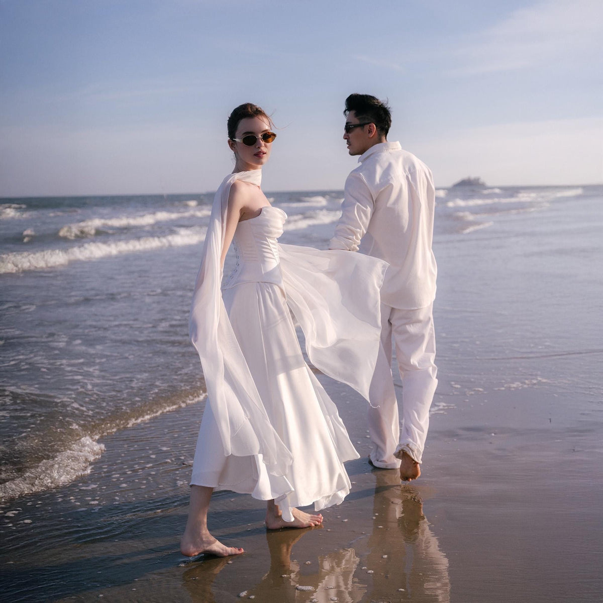 May include: A couple stands on a beach. The person on the left wears a white strapless wedding dress with a flowing skirt and a sheer cape. The person on the right wears a white suit. Both wear sunglasses.