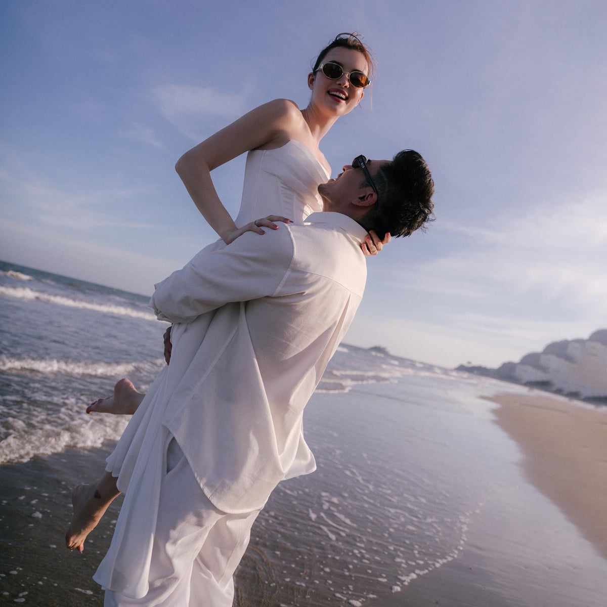 May include: A couple on a beach, the man holding the woman in his arms. Both are wearing white clothing and sunglasses. The background shows the ocean, the sky, and the beach.