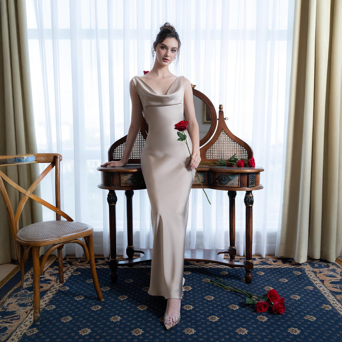 May include: A champagne-colored, floor-length gown with a cowl neckline. The dress is sleeveless and form-fitting. A red rose is held in the hand. A vintage vanity and chair are in the background. The room has a blue patterned rug and curtains.