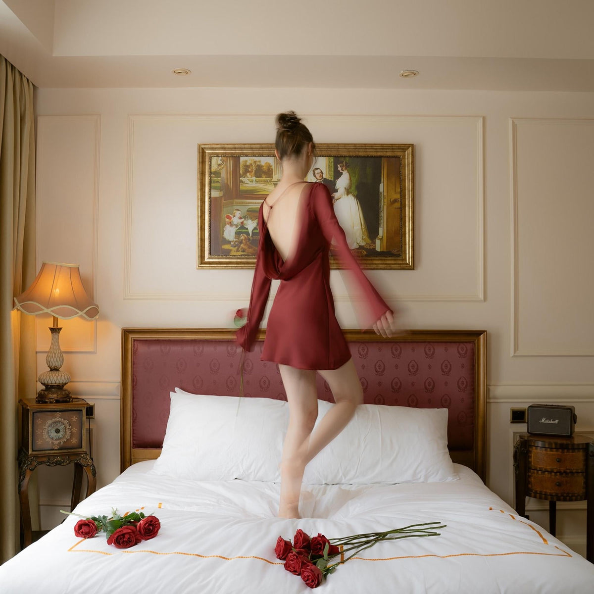 May include: A burgundy-colored dress with long sleeves and an open back. The dress is knee-length and has a draped cowl neckline. The image shows the dress in a bedroom setting, with a bed and a painting on the wall.