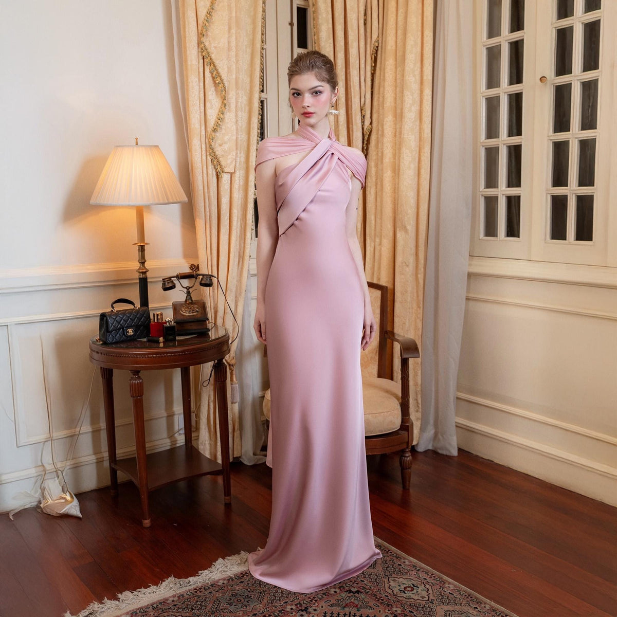 May include: A floor-length, pale pink gown with a unique halter neckline. The dress features a fitted silhouette and a subtle train. The model is standing in a room with a vintage lamp, a small table, and a window with curtains.