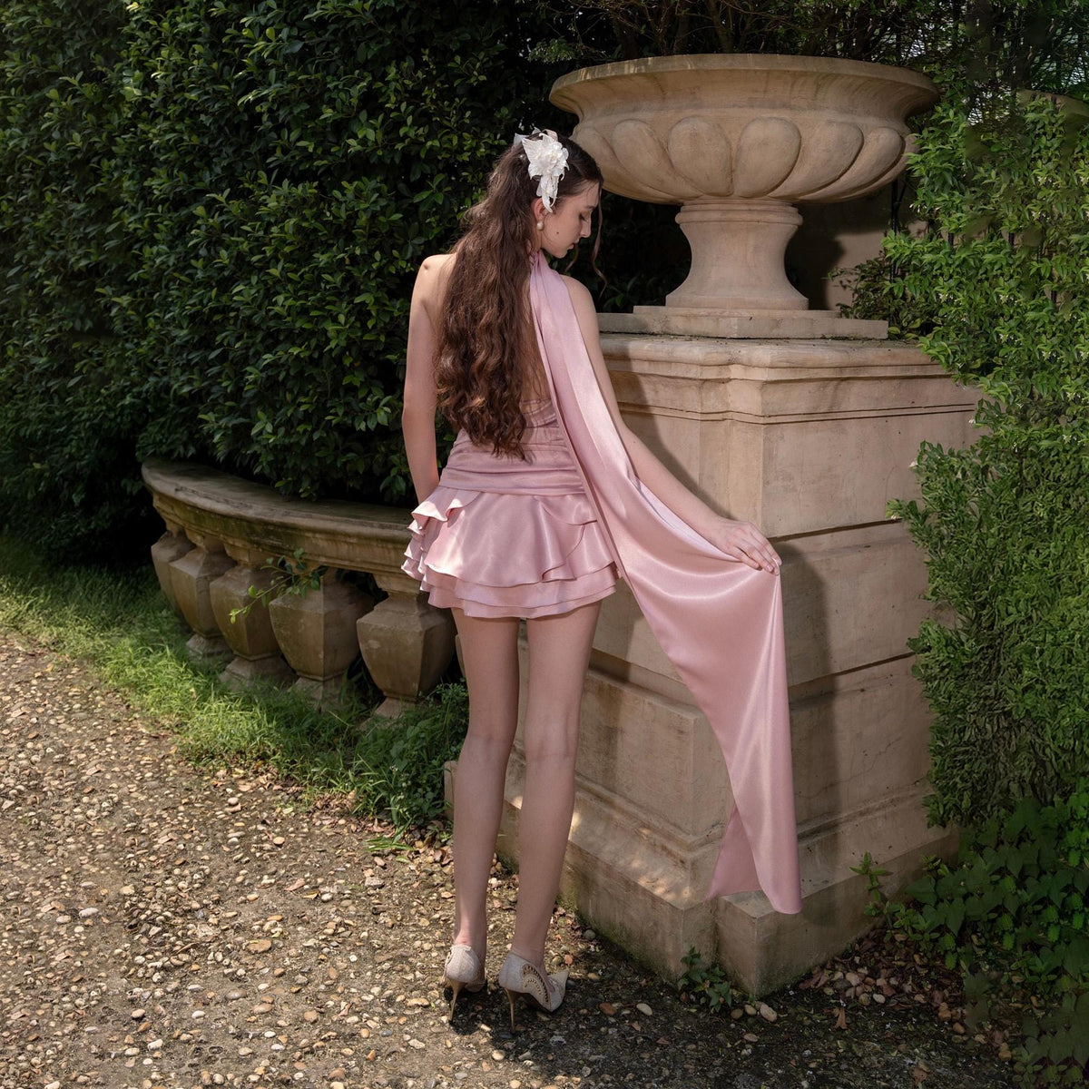 May include: A light pink satin dress with a ruffled skirt and a long, flowing scarf. The dress is paired with high-heeled shoes. The background features a stone wall, greenery, and a large decorative urn, creating an elegant outdoor setting.