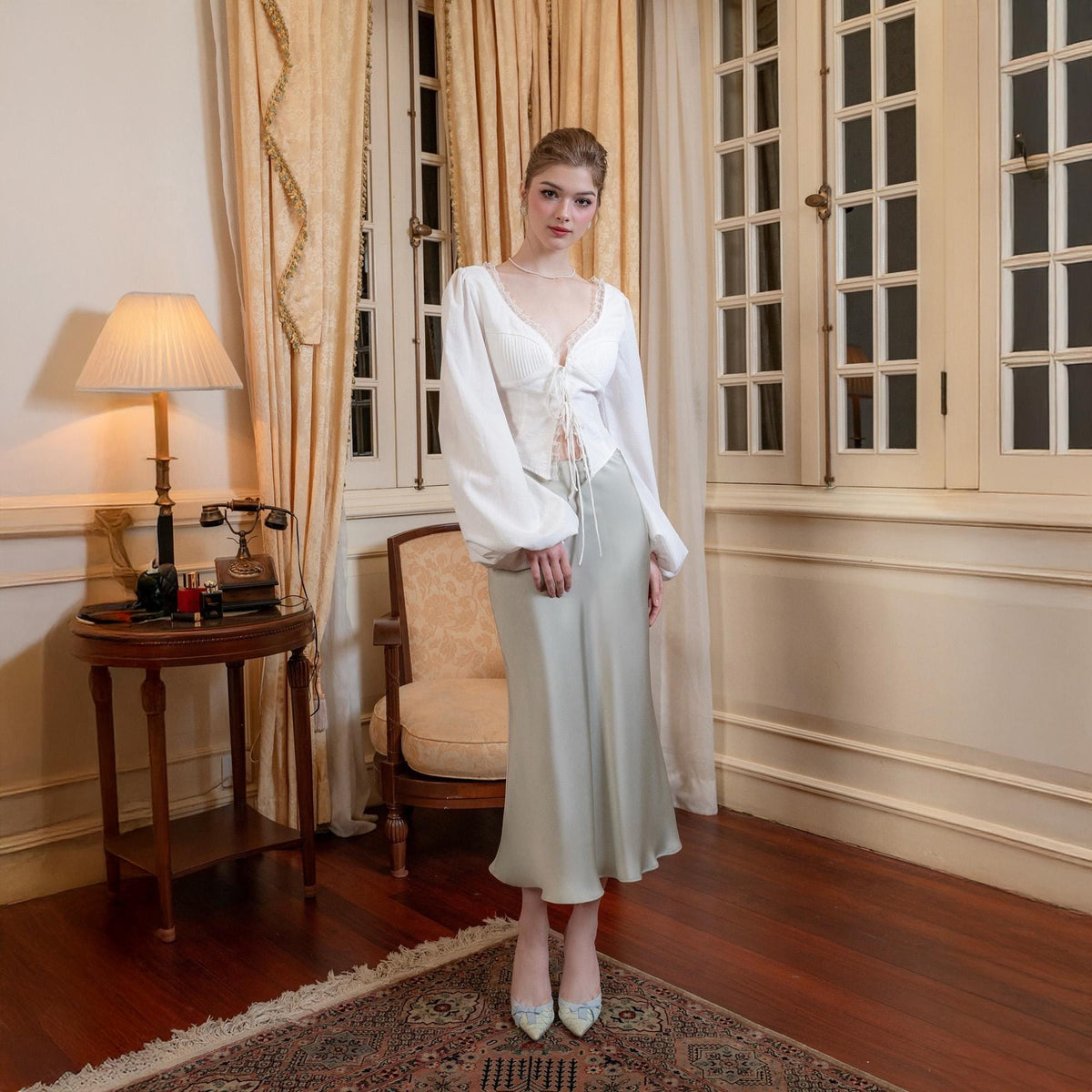 May include: A woman wearing a white blouse with long sleeves and a lace trim, paired with a light green satin skirt. The outfit is complemented by matching heels. The setting is a room with large windows and vintage decor.