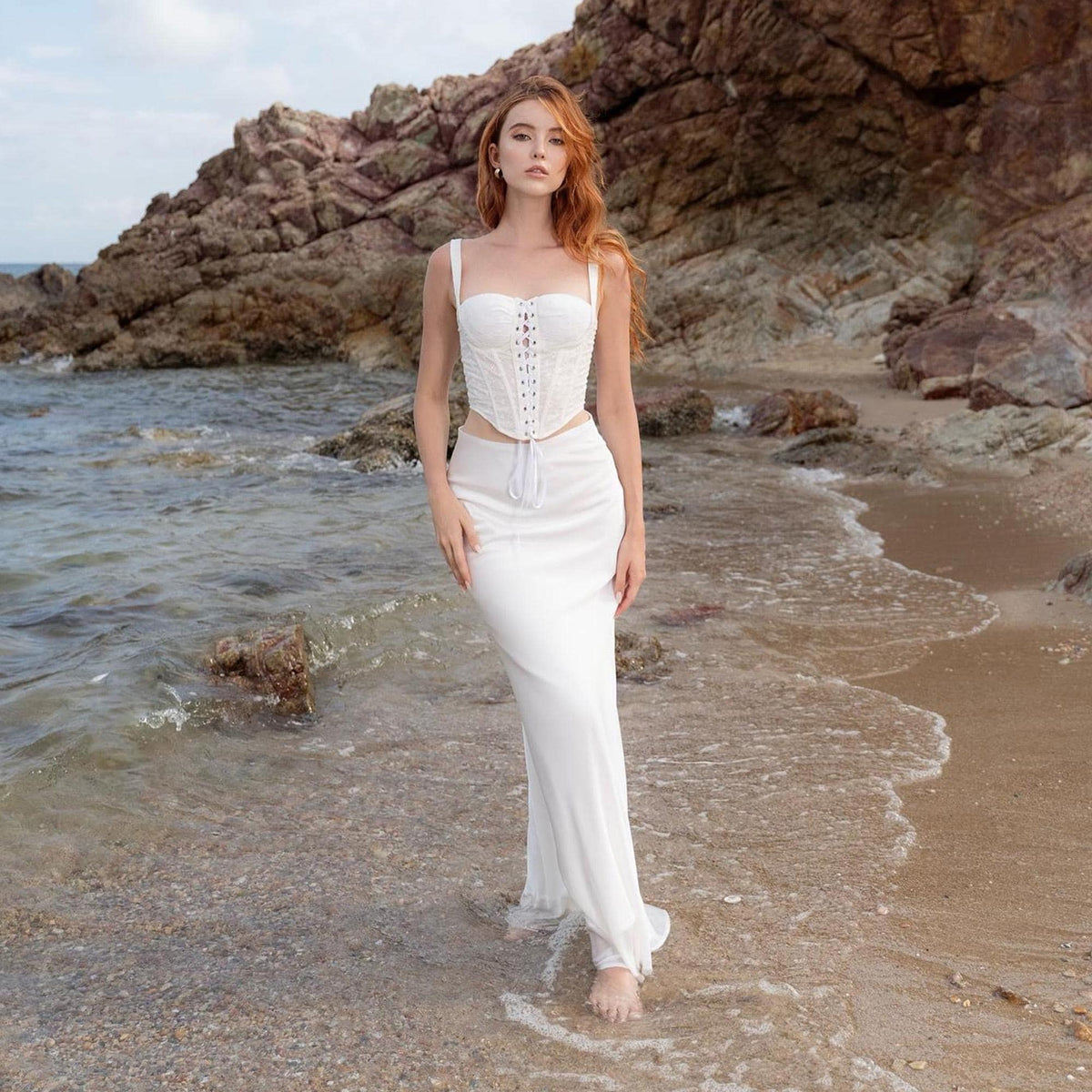 May include: A white corset top with spaghetti straps and a lace-up front, paired with a matching white skirt. The outfit is worn near the ocean, with a rocky background and the water gently lapping at the shore. The overall aesthetic is elegant and beachy.