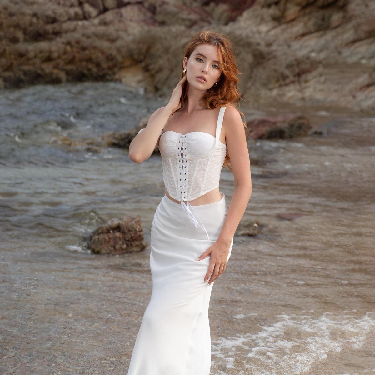 May include: A white corset top with lace detailing and spaghetti straps, paired with a matching white skirt. The outfit is worn on a beach with a rocky background and ocean waves.