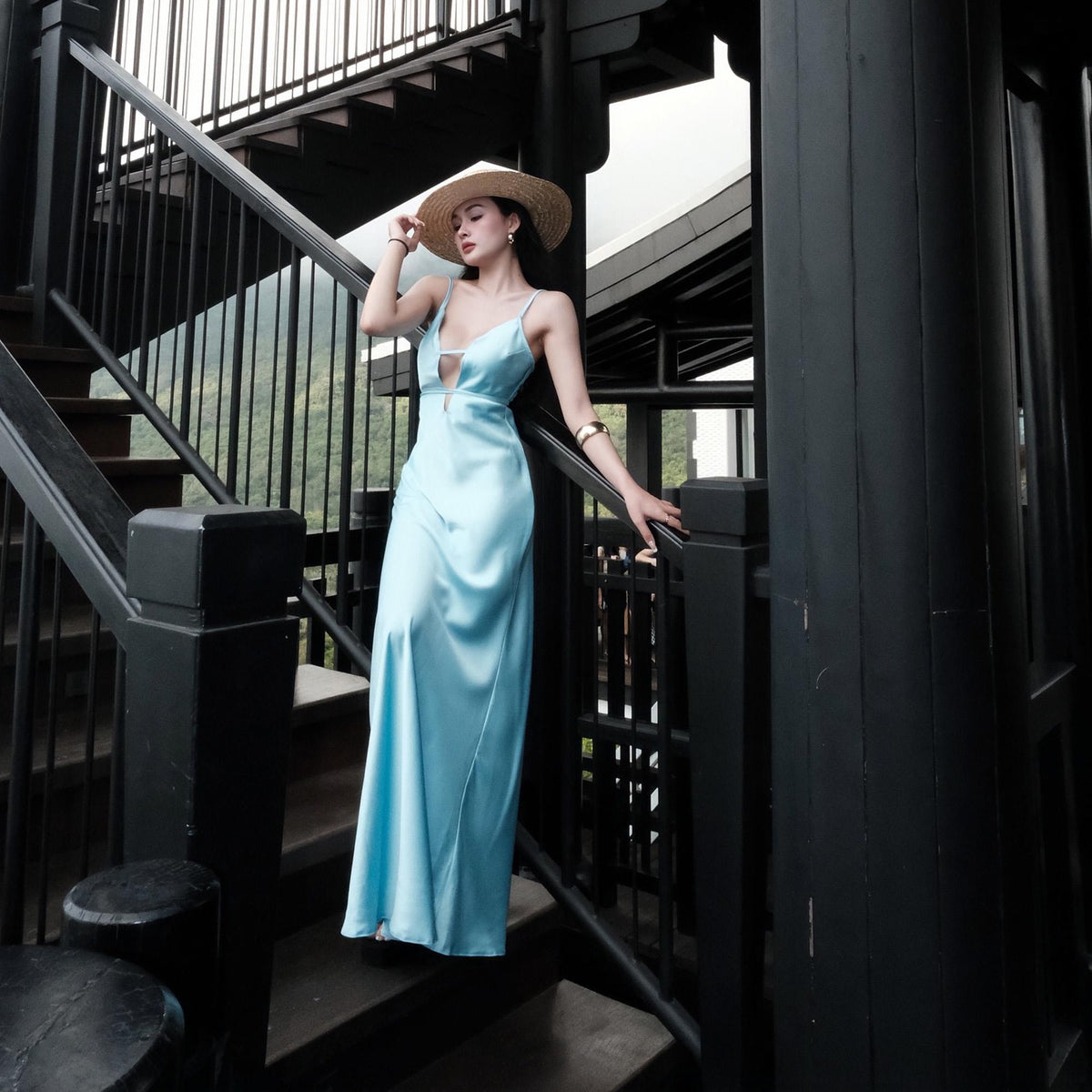 May include: A light blue satin maxi dress with a keyhole neckline. The dress features spaghetti straps and a flowing skirt. A straw hat and gold bracelet complete the look. The model is standing on a black staircase.