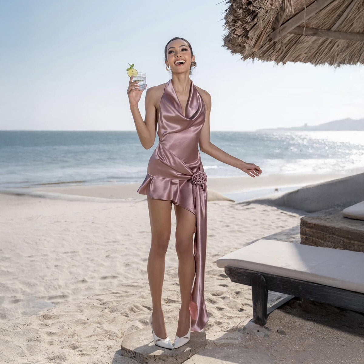 May include: A dusty rose satin halter dress with a draped cowl neckline and asymmetrical skirt. The dress features a rosette detail at the waist and a flowing side panel. The model is wearing white heels and holding a cocktail with a lime garnish.