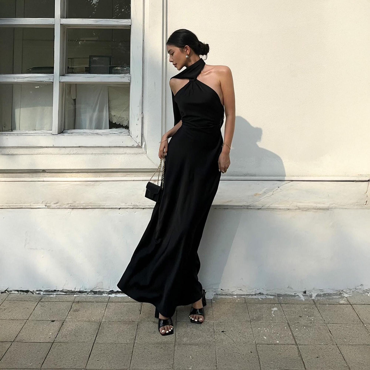 May include: A black halter neck maxi dress with a flowing skirt. The dress features a unique neckline and a single sleeve. The person is wearing black sandals and carrying a small black purse with a chain strap. The background is a white wall and a window.