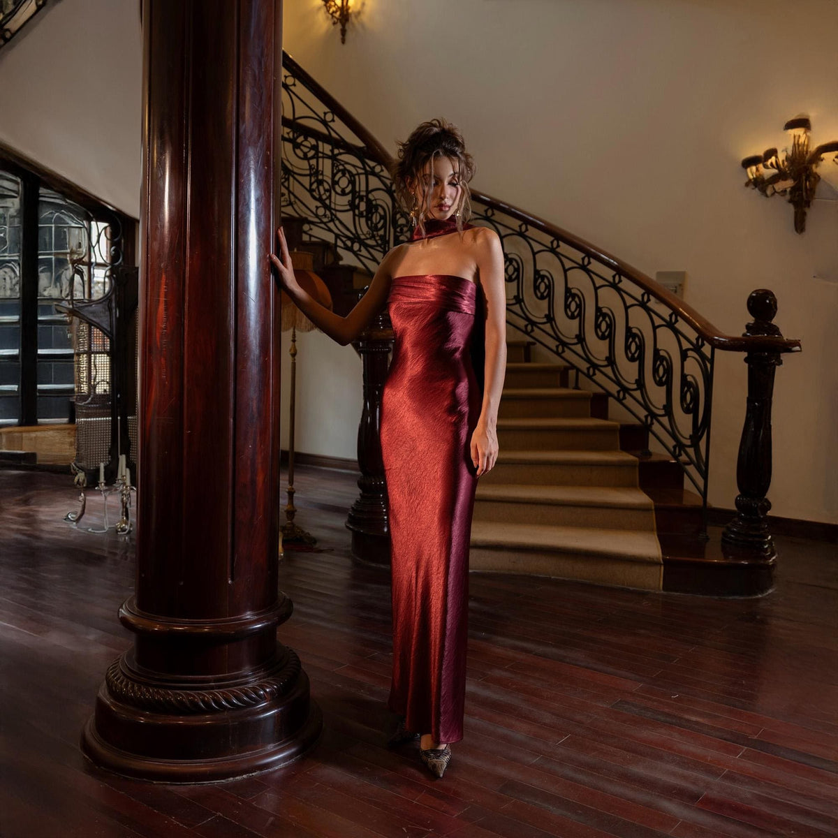 May include: A floor-length, strapless, burgundy satin gown. The dress has a fitted silhouette and a subtle sheen. The image is set in a grand interior with a wooden staircase and a large, dark wooden pillar. The dress is the focal point.