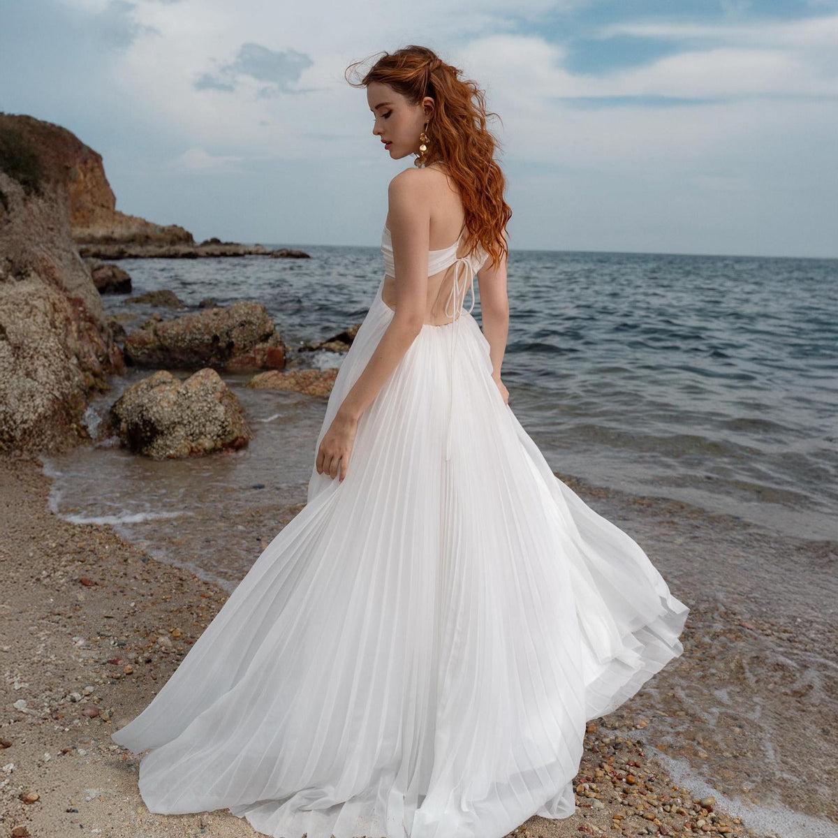 May include: A white pleated wedding dress with a halter top and open back. The dress is flowing and elegant, with a long skirt that trails on the beach. The background features the ocean and a rocky shoreline.