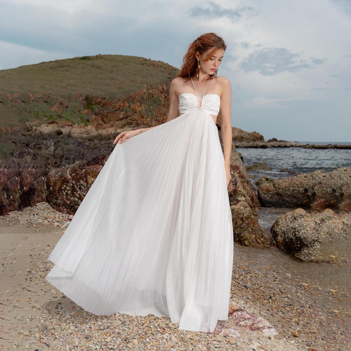 May include: A white, pleated maxi dress with a strapless bodice and cut-out detail. The dress is flowing and elegant, perfect for a beach or outdoor event. The model is standing on a sandy beach with rocks and the ocean in the background.