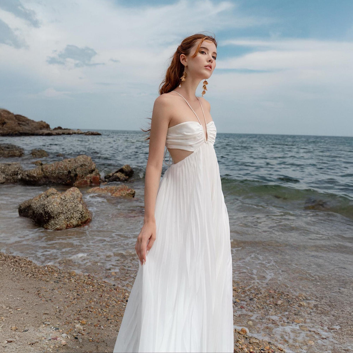 May include: A white, floor-length dress with a halter neckline and pleated skirt. The dress features cut-out details at the waist. The photo is taken on a beach with the ocean and rocks in the background.