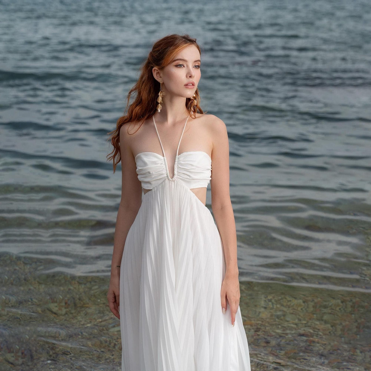 May include: A white halter dress with a pleated skirt and cut-out detail. The dress has a draped bodice and a delicate halter strap. The model is standing near the water, with the ocean in the background. The dress is a formal or semi-formal style.