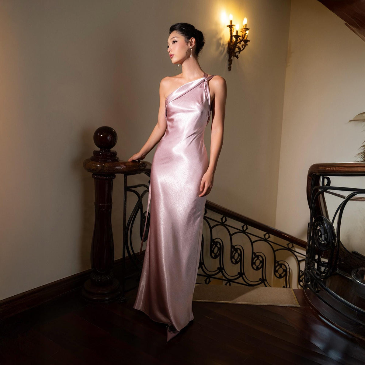 May include: A light pink, one-shoulder satin gown. The dress has a flowing, floor-length design. The model is standing on a wooden staircase with a dark brown handrail. The background features a wall with a sconce.