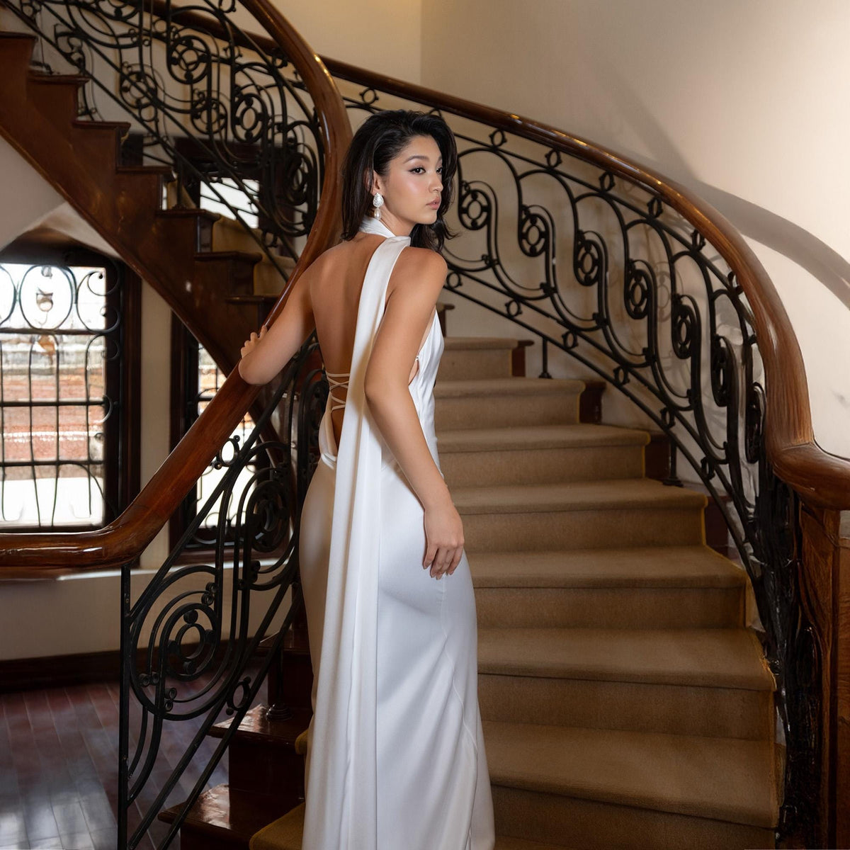May include: A white, floor-length gown with a draped back and halter neckline. The dress is worn on a staircase with a dark wood banister and ornate wrought iron railing. The stairs are covered in a neutral-toned carpet.