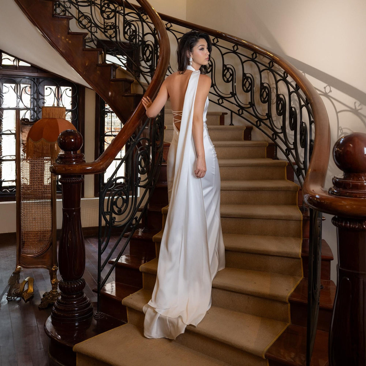 May include: A white satin gown with a flowing train. The dress features a halter neckline and a low back with crisscross straps. The model is standing on a wooden staircase with a decorative wrought iron railing. The stairs are covered in a beige carpet.