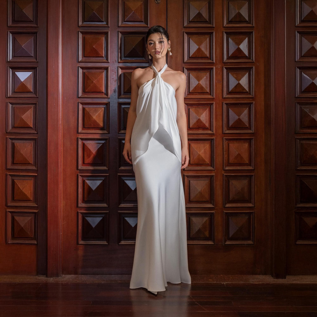 May include: A floor-length, white halter dress with a flowing skirt. The dress features a draped bodice and a simple, elegant design. The model is standing in front of a dark wooden door.