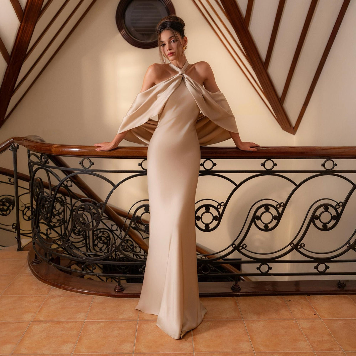 May include: A beige, floor-length gown with a halter neckline and attached cape-like sleeves. The dress is made of a smooth, flowing fabric. The model is leaning on a dark wooden railing with decorative metalwork.