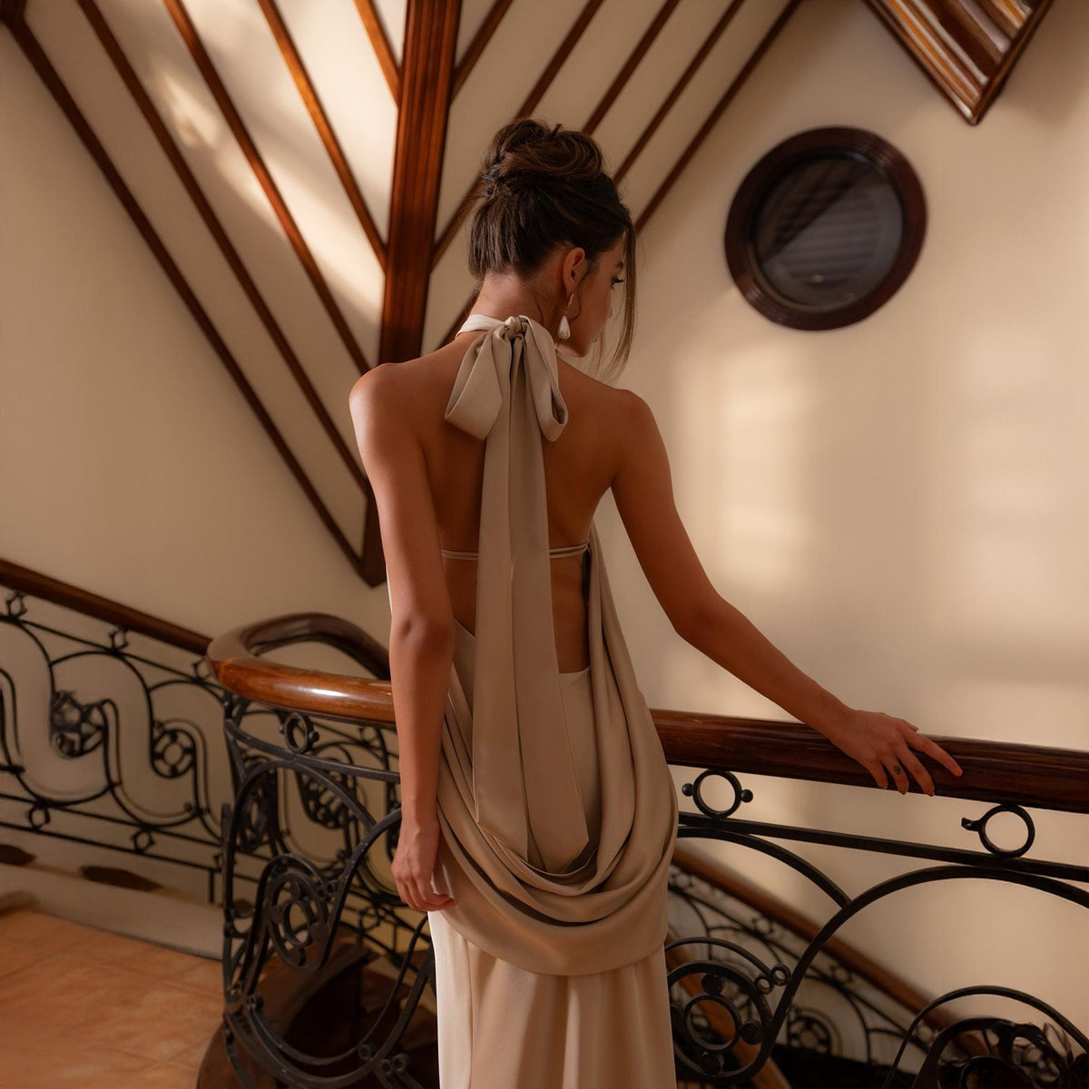 May include: A beige halter-neck gown with a draped back detail. The dress has a flowing skirt and a bow at the neck. The model is standing on a staircase with a wooden banister and wrought iron railing. The overall aesthetic is elegant and sophisticated.