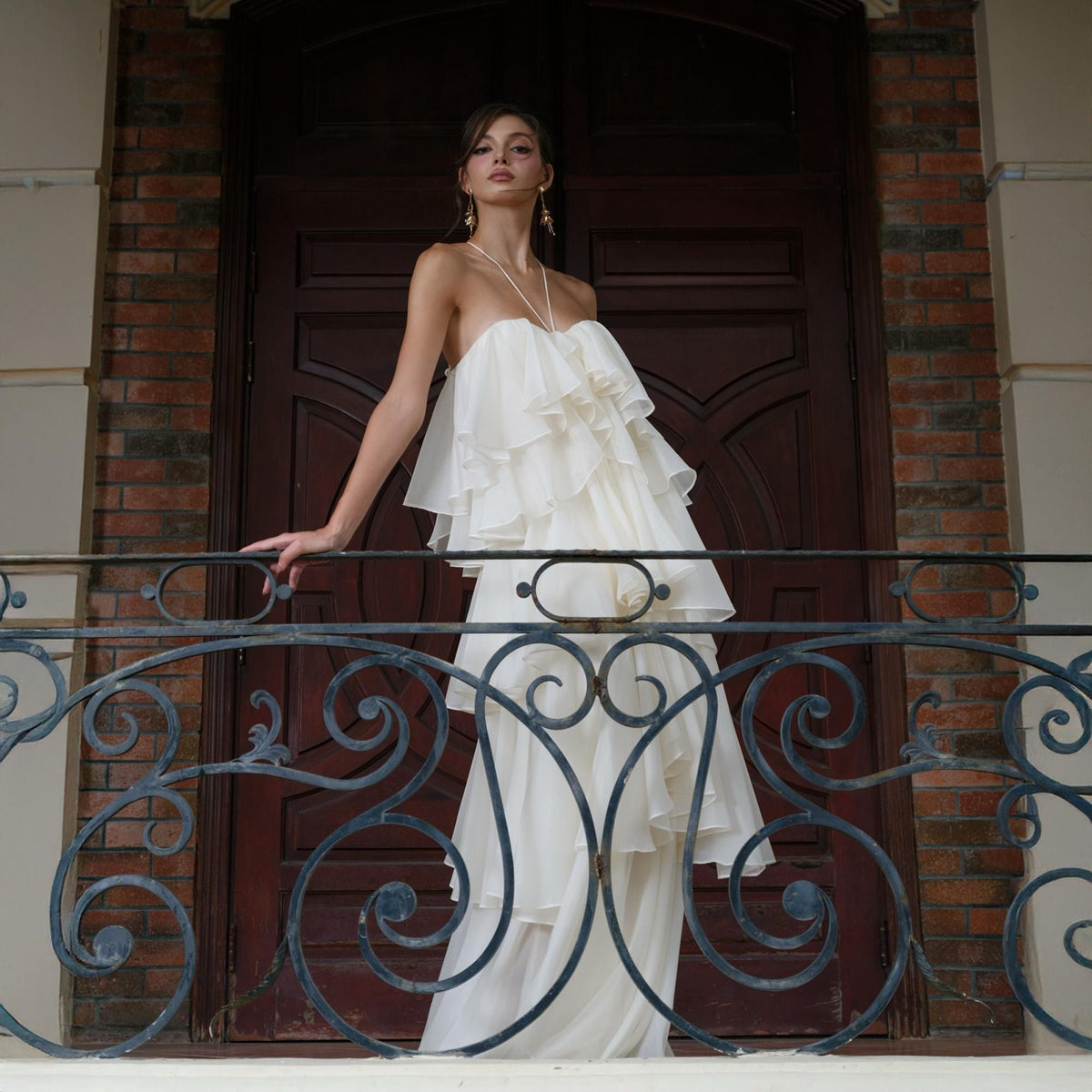 May include: A strapless, ivory-colored gown with layered ruffles. The dress has a flowing skirt and is worn with delicate jewelry. The background features a dark wooden door and a wrought iron balcony.
