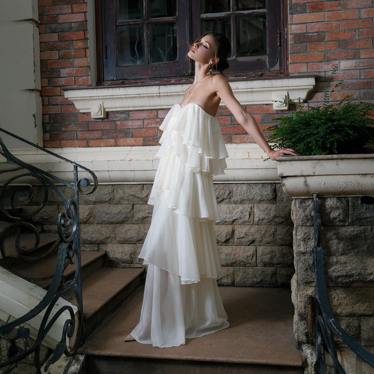 May include: A strapless, ivory-colored gown with layered ruffles. The dress cascades down the body, creating a flowing silhouette. The model is standing on outdoor steps, with a brick building in the background. The dress is suitable for a wedding or formal event.