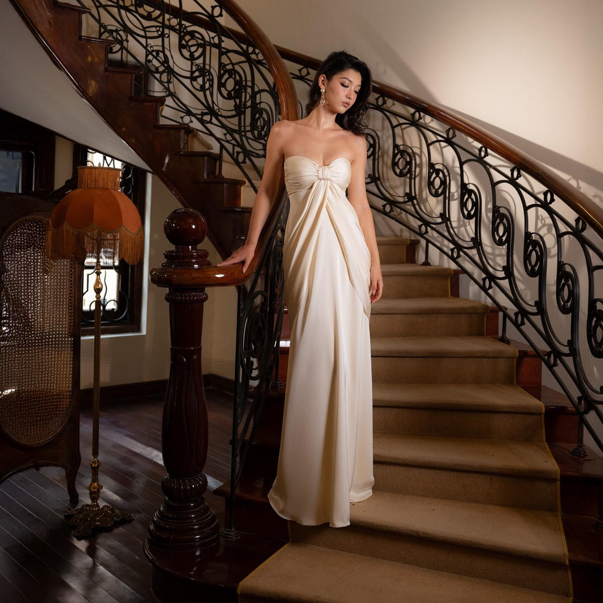 May include: A strapless, ivory-colored gown with a draped bodice and flowing skirt. The dress is worn on a staircase with a dark wooden banister and ornate wrought iron railing. The setting is a well-lit interior.