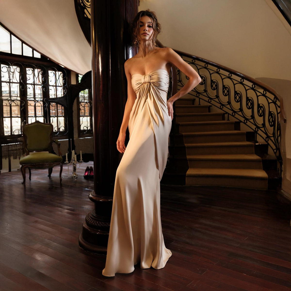 May include: A strapless, floor-length champagne-colored gown with a draped bodice. The dress features a fitted silhouette and a subtle train. The model is standing in a grand, well-lit interior with a spiral staircase and dark wood flooring.