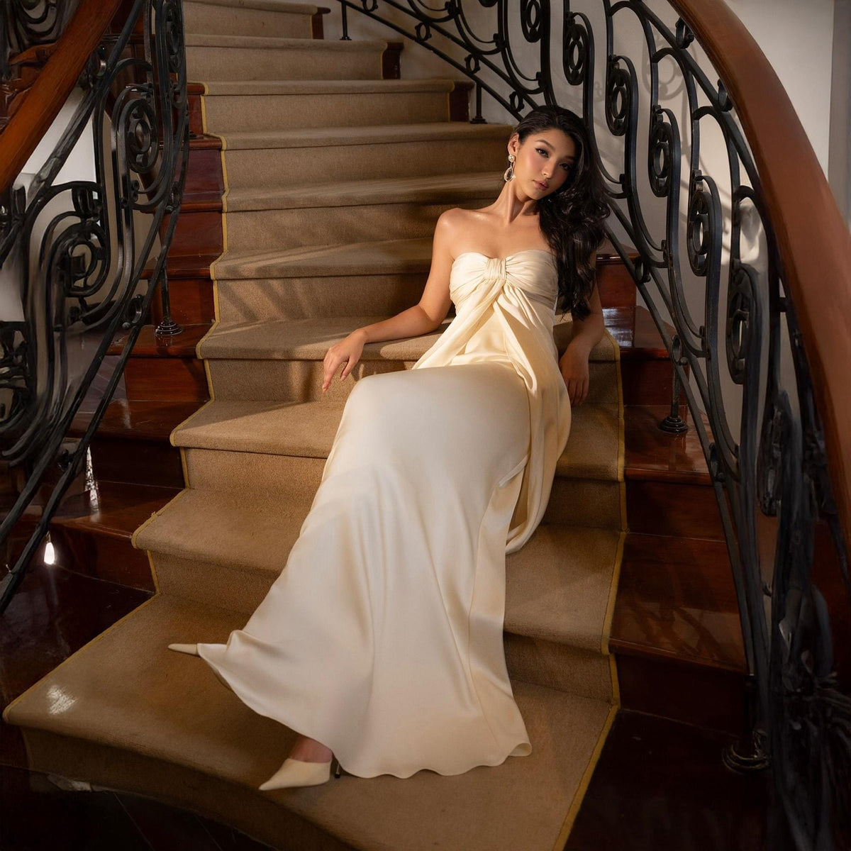 May include: A strapless, floor-length ivory gown with a draped bodice and flowing skirt. The dress is worn on a staircase with a brown wooden banister and ornate black metal railing. The stairs are covered in a neutral-toned carpet.