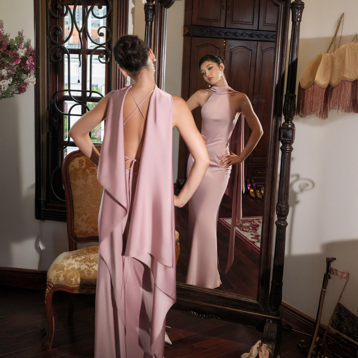 May include: A dusty rose-colored formal gown with a halter neckline and a flowing cape detail. The dress is floor-length and has a fitted silhouette. The back of the dress features a crisscross strap design. The image is taken in front of a large mirror.