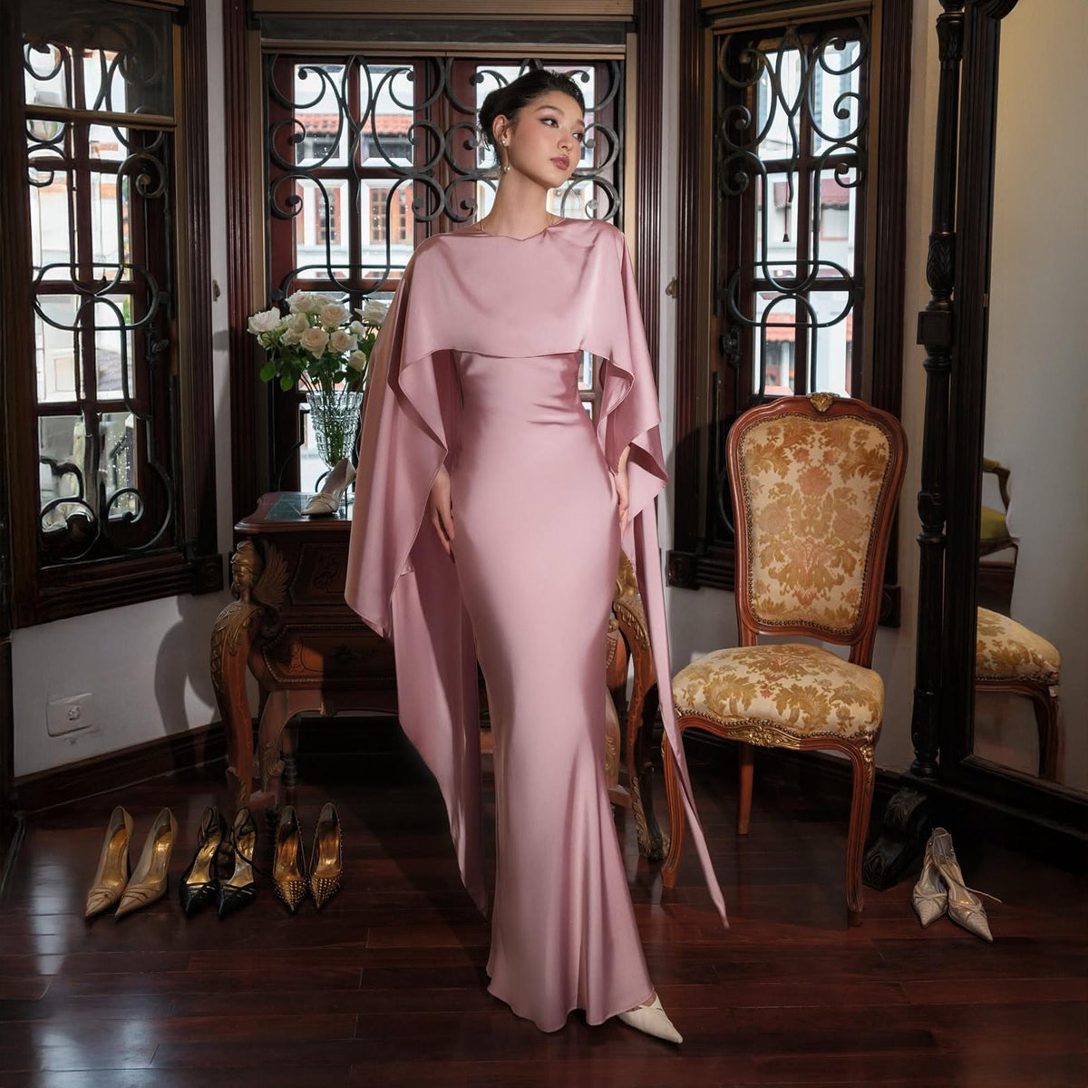 May include: A floor-length, pale pink gown with a cape-like overlay. The dress has a fitted silhouette and a flowing cape that drapes over the shoulders and arms. Several pairs of high heels in gold and black are on the floor. The setting is a room with antique furniture.