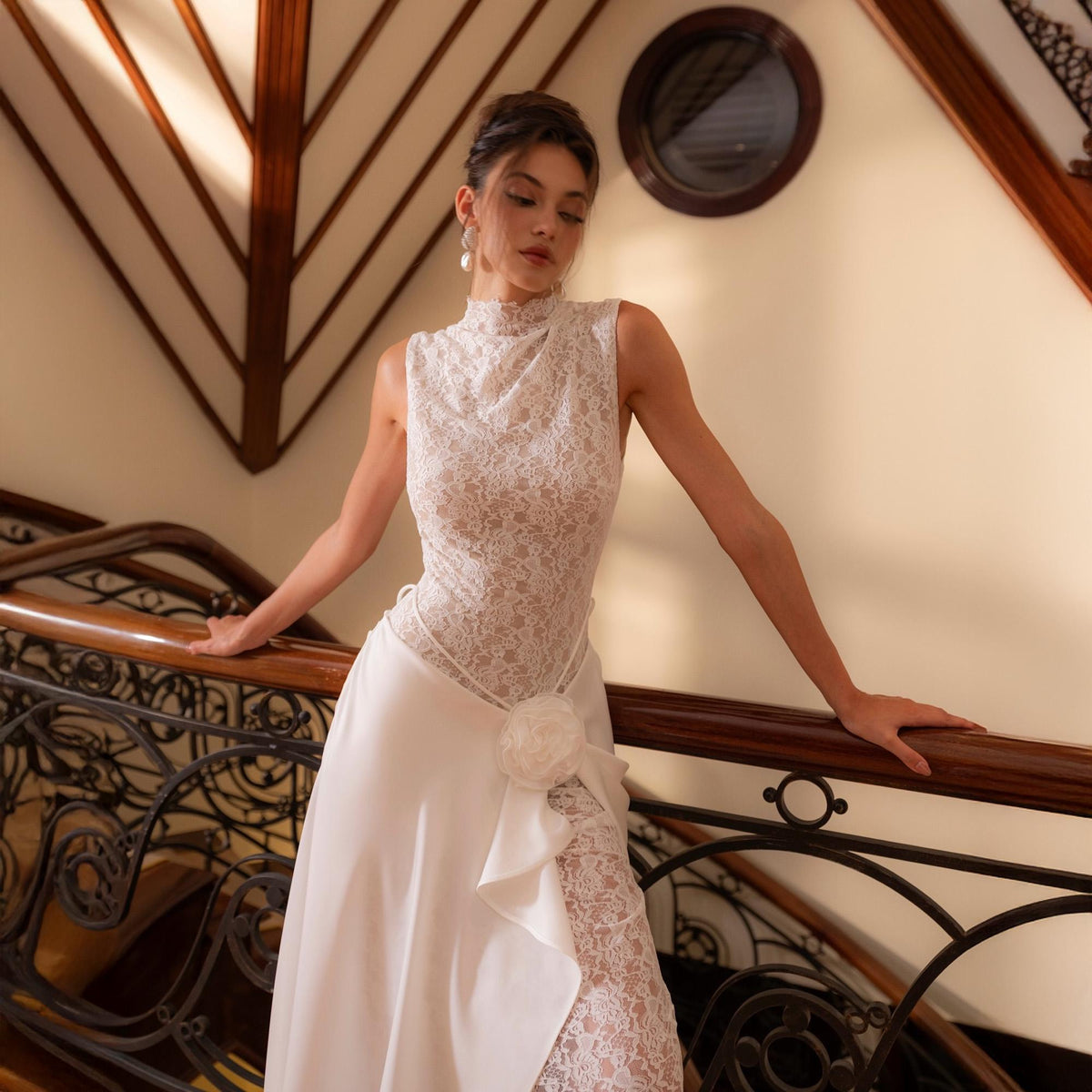 May include: A white sleeveless wedding dress with a high neckline and a lace bodice. The skirt is flowing, with a floral accent at the waist. The dress is worn on a wooden staircase with ornate metal railings.