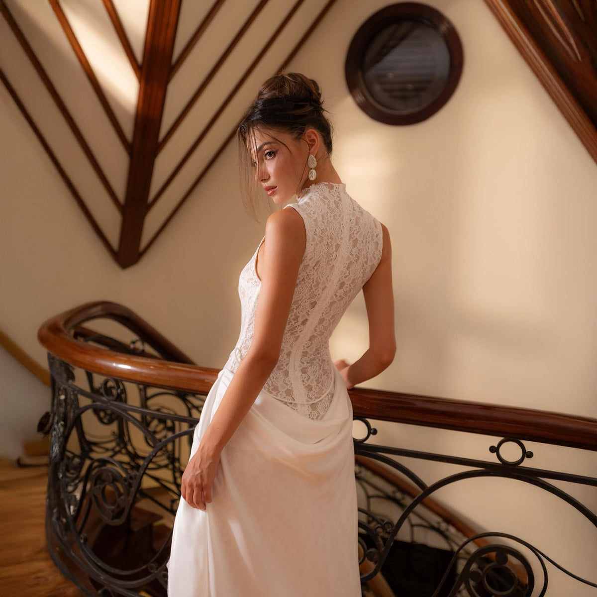 May include: A sleeveless white lace bodice wedding dress with a high neckline and a flowing skirt. The dress is worn on a wooden staircase with a decorative railing. The background includes a round window and architectural details.