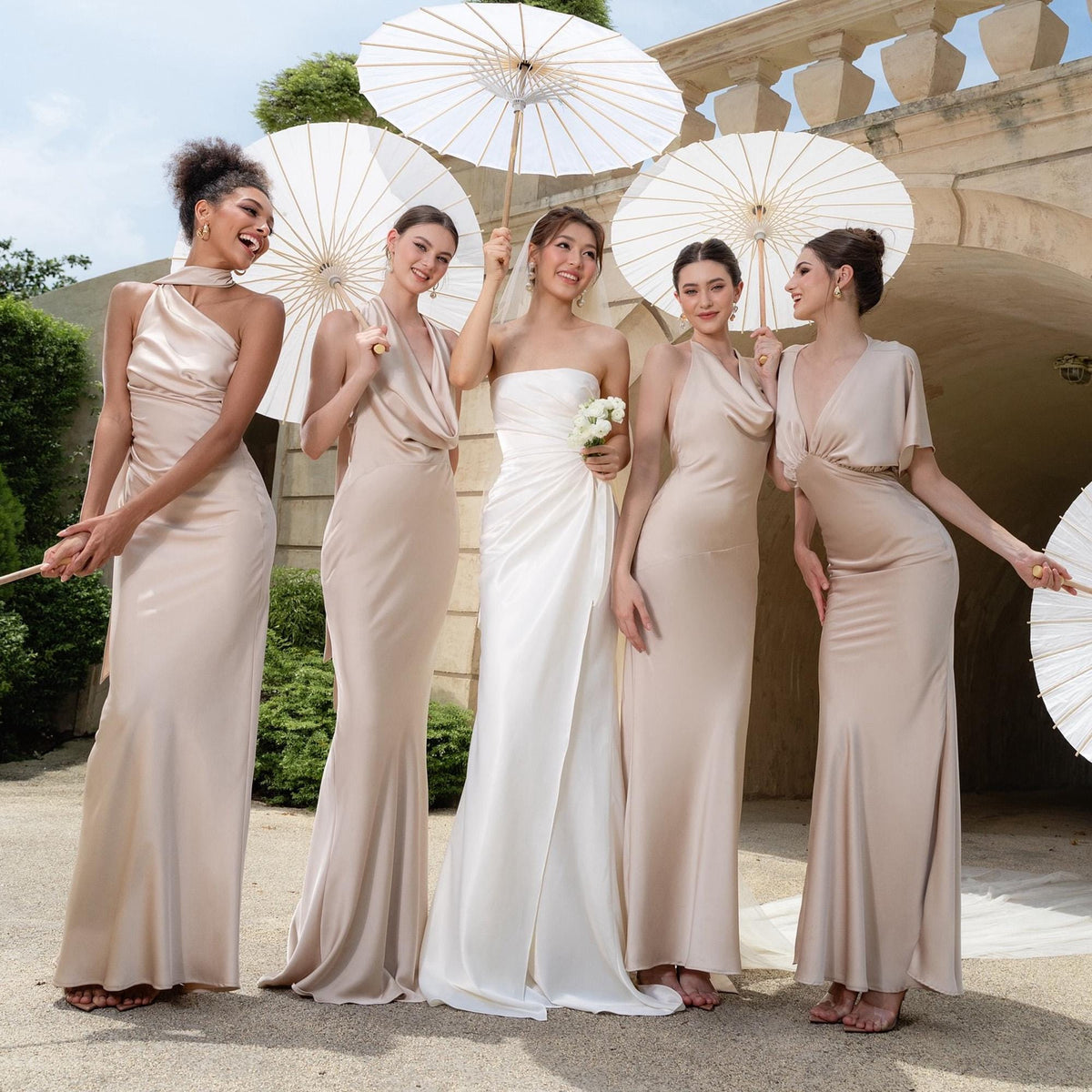 May include: A bride in a white strapless gown stands with four women in champagne-colored dresses. Each woman holds a white parasol. The group is outdoors, posing in front of a stone structure. The bride holds a small bouquet of white flowers.