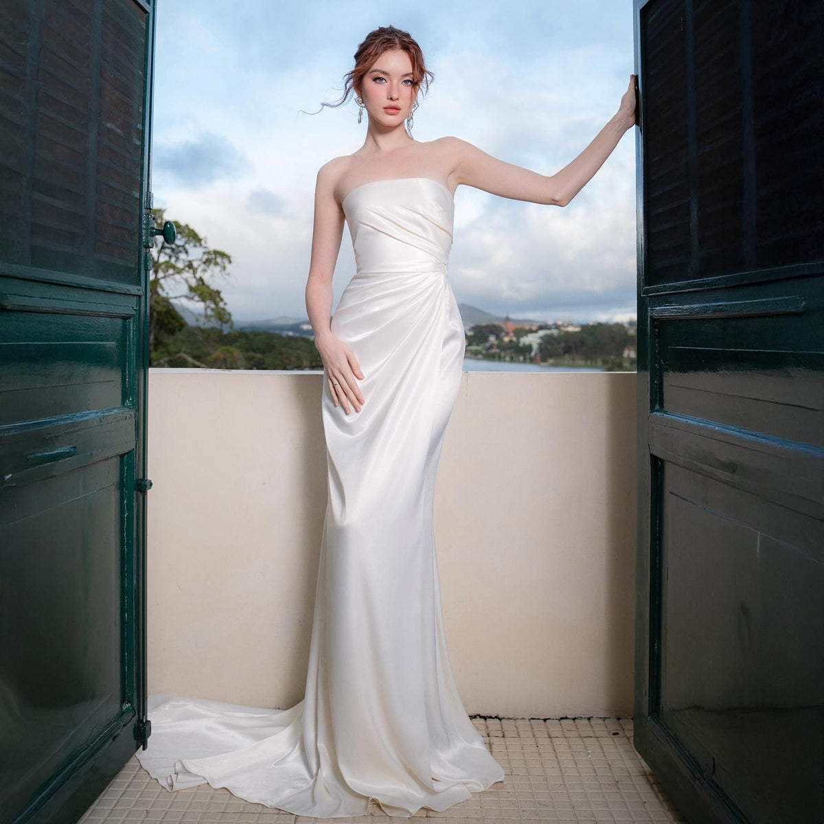 May include: A strapless white satin gown with a draped bodice and a flowing skirt. The dress features a subtle train and is worn by a person standing on a balcony with a scenic view in the background. The door frame is dark green.