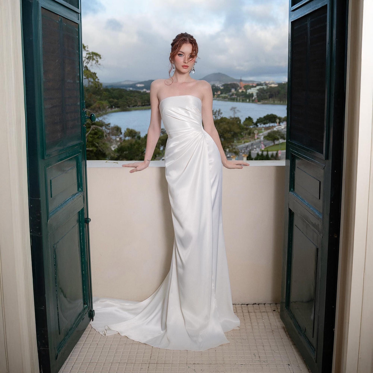 May include: A strapless white satin gown with a draped bodice and flowing skirt. The dress features a subtle train and is worn by a person standing on a balcony overlooking a scenic landscape.