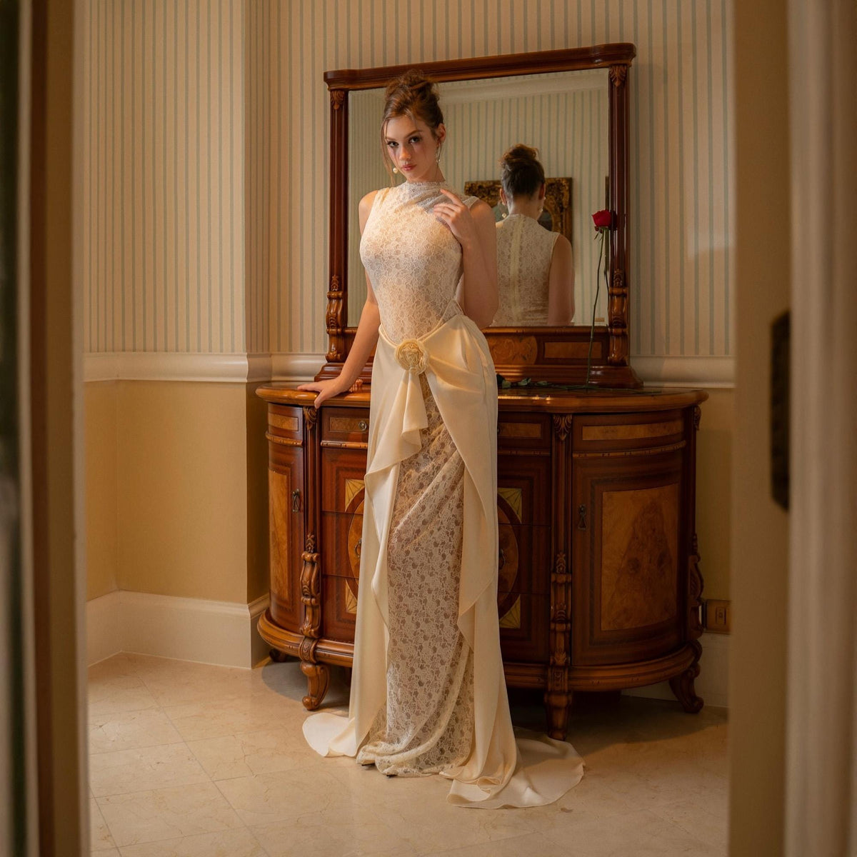May include: A cream-colored formal gown with a high neckline and floral lace detailing. The dress features a draped skirt with a bow at the waist. A large wooden dresser with a mirror is in the background.