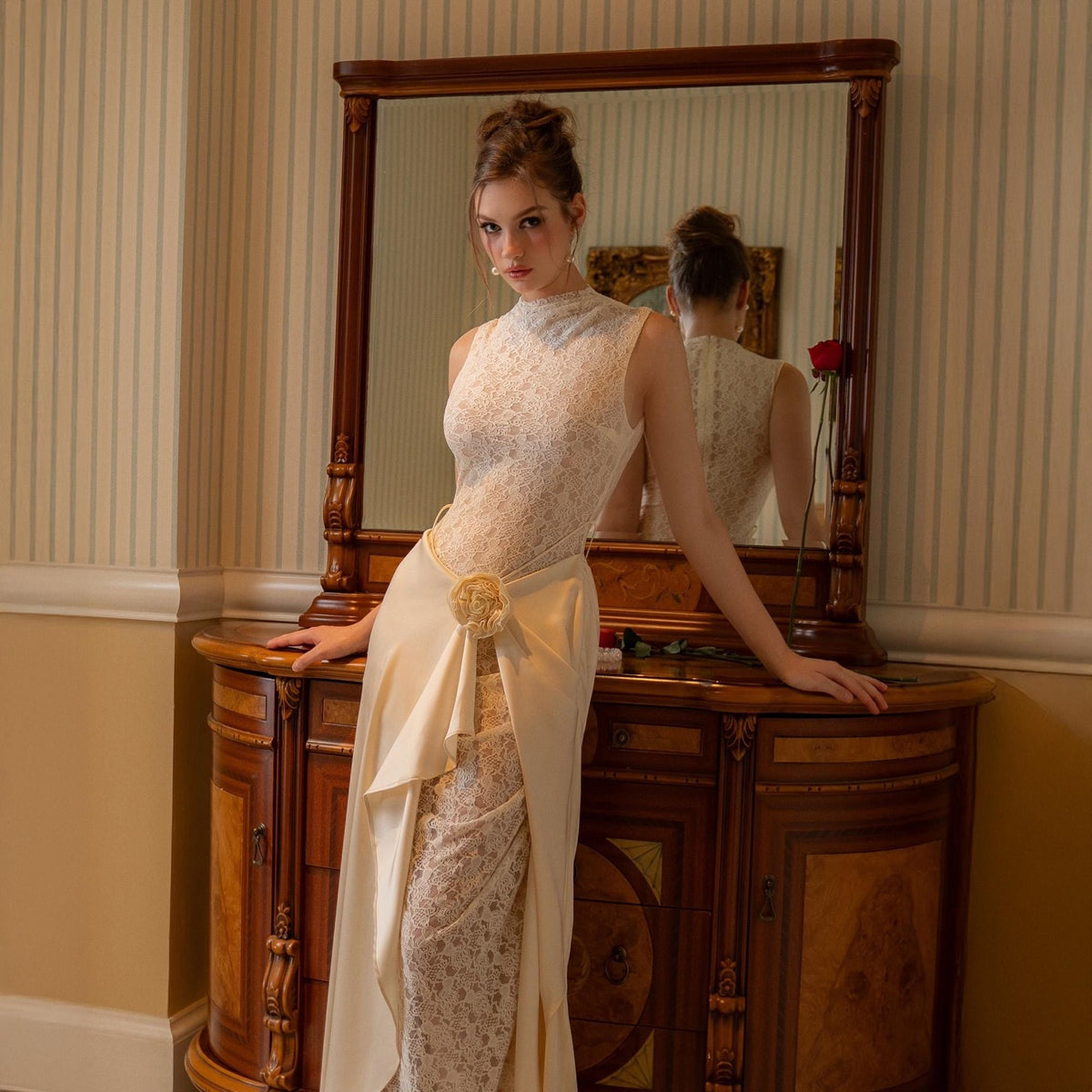 May include: A sleeveless ivory lace gown with a high neckline and a flowing skirt. The dress features a satin overlay with a decorative floral accent at the waist. A red rose rests on a wooden dresser.