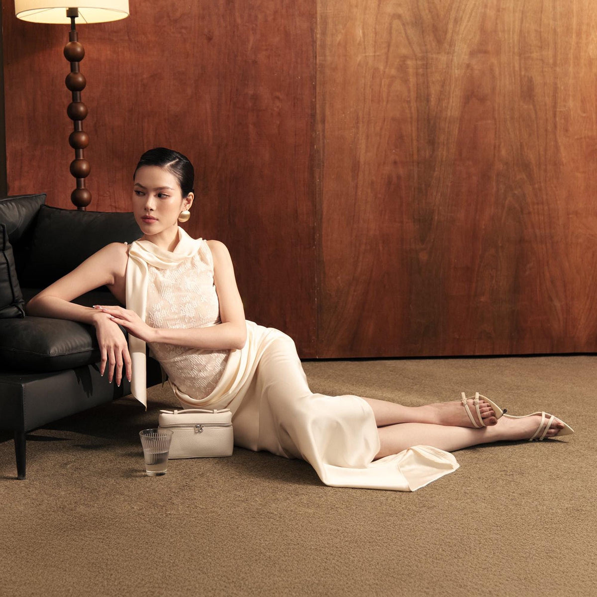 May include: A woman wearing a cream-colored dress with a high neckline and a fitted bodice. The dress is paired with strappy sandals. A small white handbag and a glass of water are nearby. The background features a dark brown wooden wall and a black sofa.