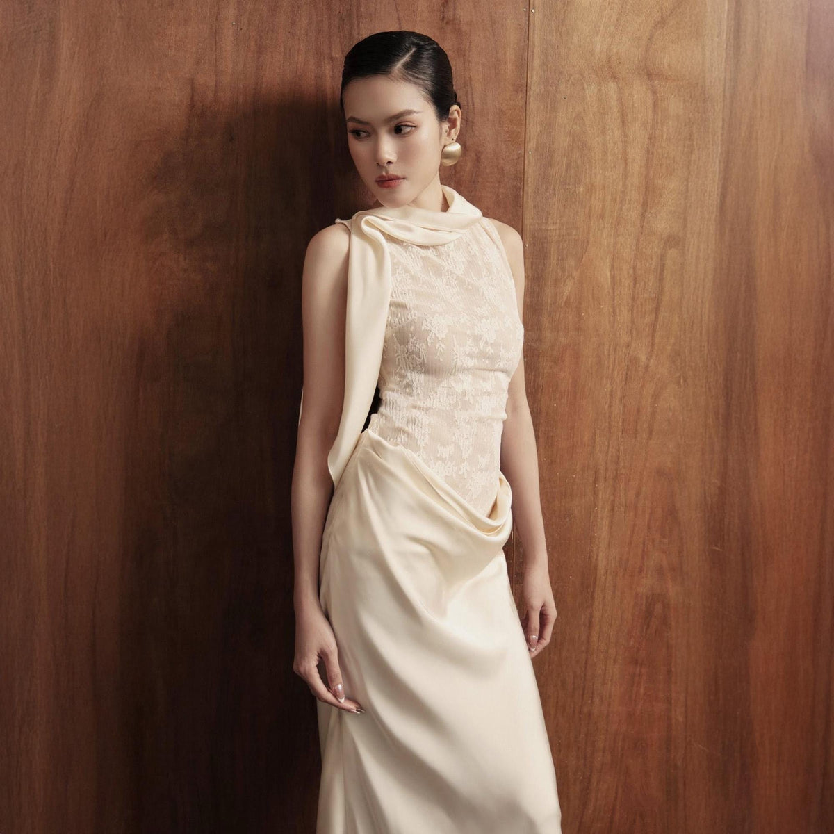 May include: A sleeveless, ivory-colored dress with a draped design. The bodice features a lace overlay, and a scarf-like element drapes over the shoulder. The dress has a flowing, floor-length skirt, creating an elegant silhouette.