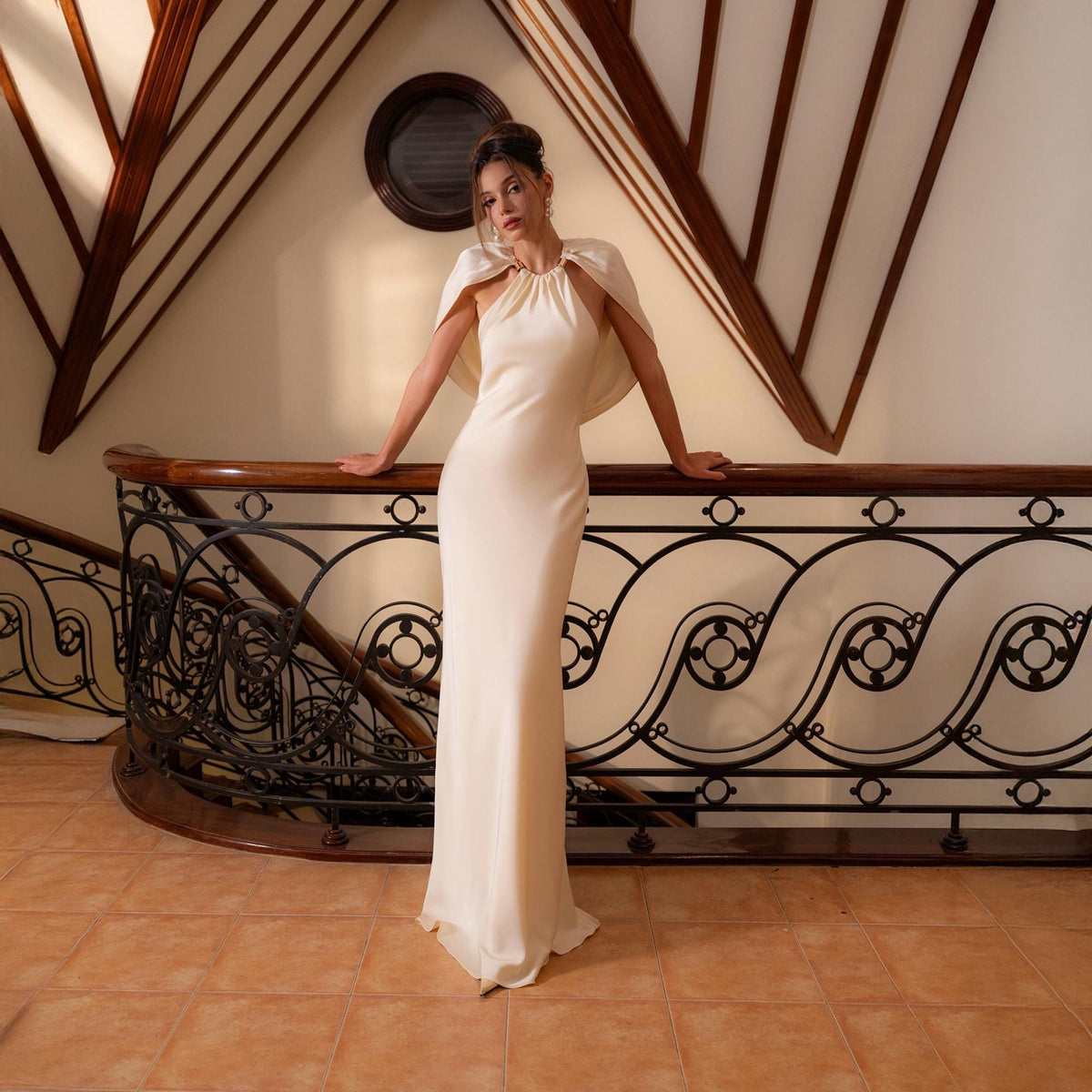 May include: A cream-colored, floor-length gown with a halter neckline and a matching cape. The dress is form-fitting and flows to the floor. The model is standing on a staircase with a decorative wrought iron railing.