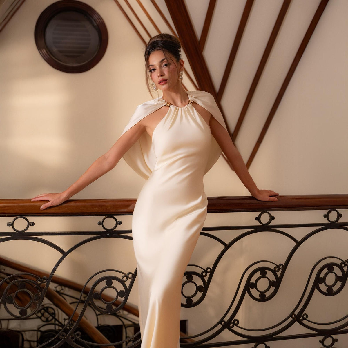 May include: A cream-colored, floor-length gown with a halter neckline and a matching cape. The dress has a simple, elegant design. The model is leaning on a dark wooden railing with decorative metalwork.