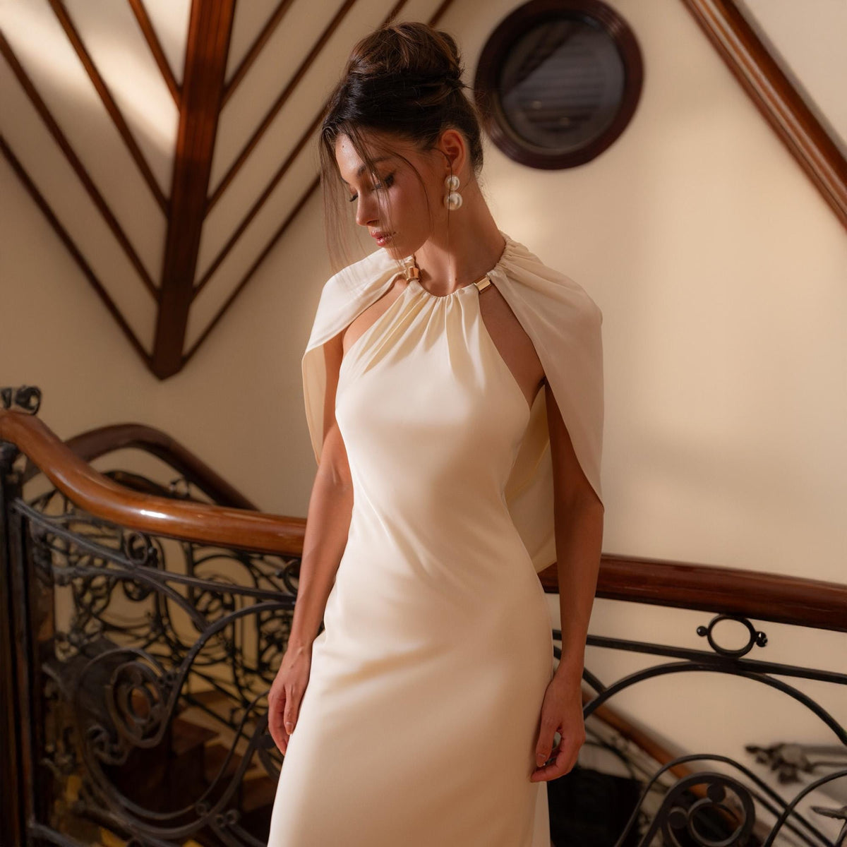 May include: An ivory-colored halter dress with a matching cape. The dress has a simple, elegant design, with a fitted bodice and a flowing skirt. The cape is draped over the shoulders and is secured at the neckline with a gold-colored clasp. The dress is suitable for formal events.