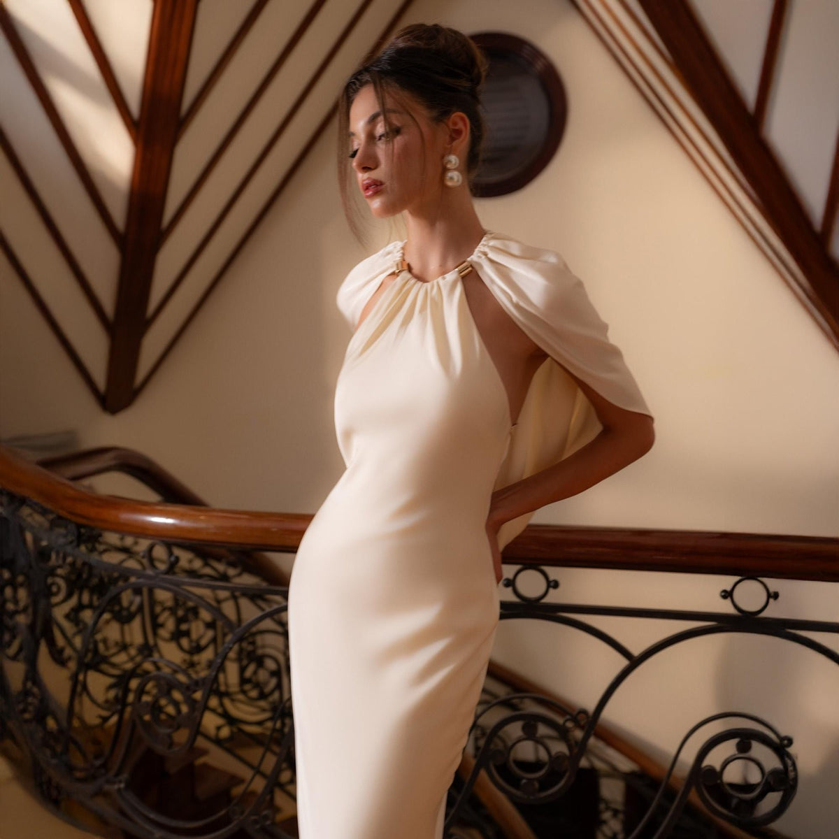 May include: A cream-colored, floor-length gown with a halter neckline and a cape-like overlay. The dress features a fitted silhouette and is accessorized with pearl earrings. The background includes a wooden staircase with ornate metal railings.