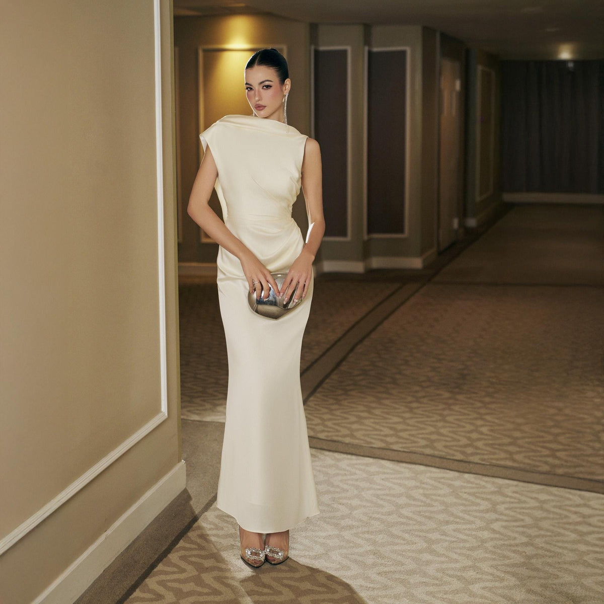 May include: A woman in a cream-colored, floor-length gown with a draped neckline and a fitted silhouette. She holds a silver clutch. The gown is paired with silver heels. The background is a hallway with neutral-toned walls and carpet.